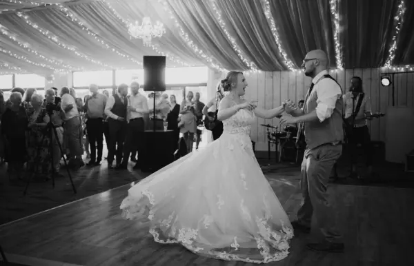 A couple dances joyfully on a wooden floor, surrounded by guests in a beautifully draped hall adorned with twinkling lights.