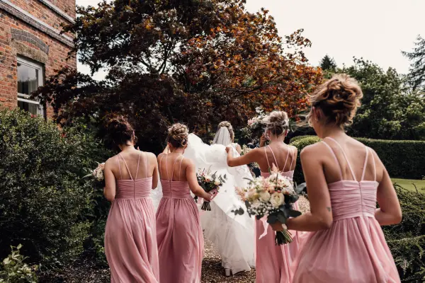 A bride in a flowing white gown walks down a lush path with her bridesmaids in soft pink dresses amidst vibrant greenery and trees.