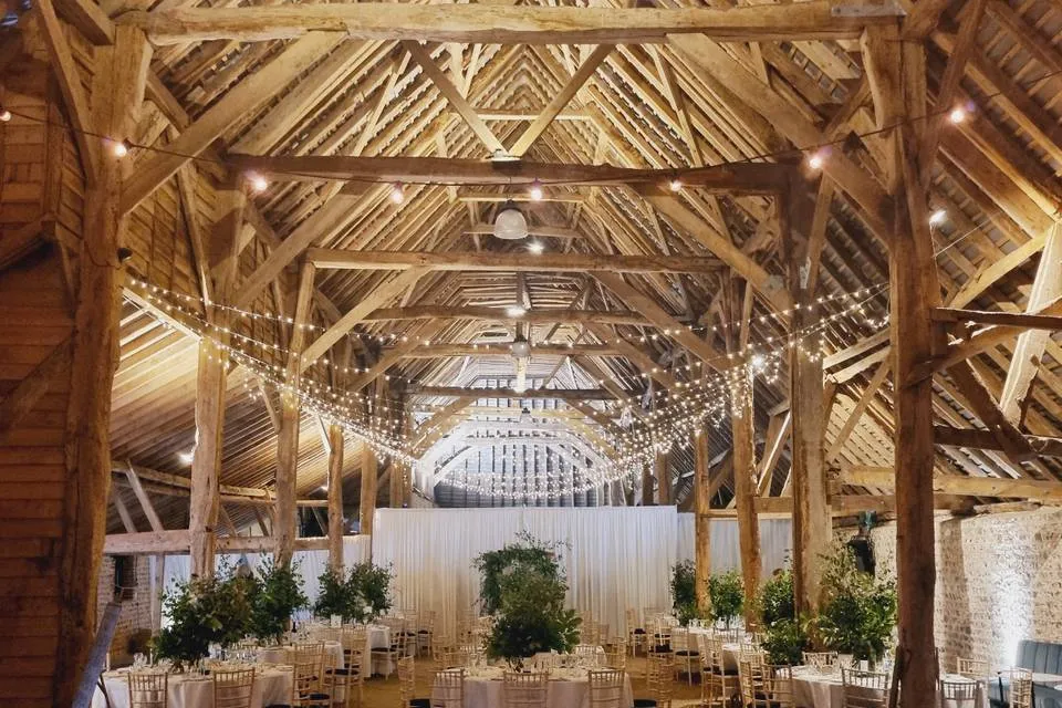 Rustic barn venue featuring high wooden beams, string lights, and elegant round tables surrounded by lush greenery for a charming wedding setting.