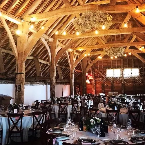 Charming rustic barn interior featuring exposed wooden beams, elegant tables set for a wedding, and warm ambient lighting.