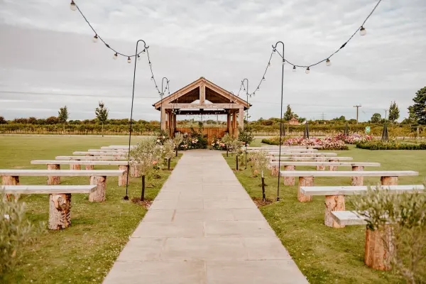 Charming outdoor ceremony space featuring a rustic wooden arch, log seating, and string lights in a beautifully landscaped setting.
