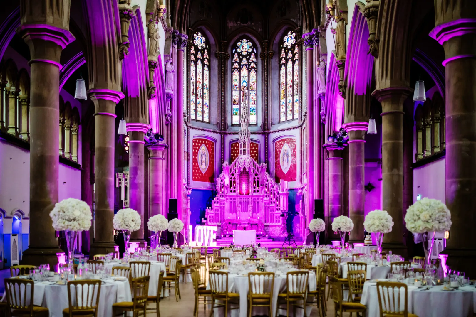Elegant wedding venue featuring a stunning stained glass backdrop, ornate architecture, and vibrant pink lighting with round tables adorned with floral centerpieces.