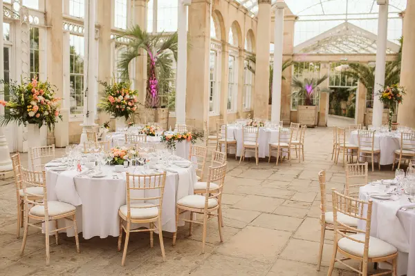 Bright glasshouse with elegant round tables, Chiavari chairs, and vibrant floral centerpieces, surrounded by lush greenery and natural light.