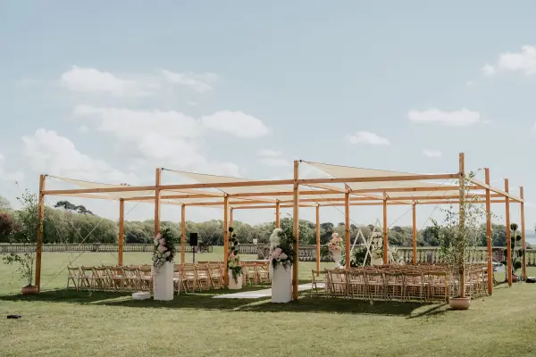An elegant outdoor ceremony space featuring a wooden pergola, draped fabric, and stylish seating surrounded by greenery.