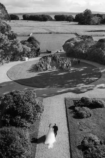 A couple walks towards a circular ceremony space surrounded by greenery, with serene water and a scenic landscape in the background.