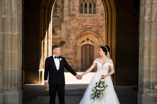 A couple stands hand-in-hand under a grand stone archway, showcasing intricate architectural details, perfect for a romantic wedding setting.