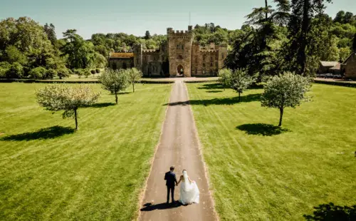 A newlywed couple walks hand in hand down a lush, tree-lined path leading to a grand castle surrounded by scenic greenery.