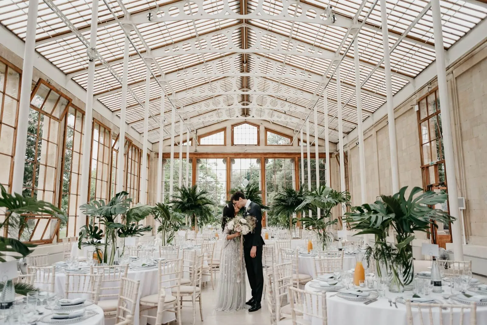 A stunning glass conservatory adorned with lush greenery, featuring elegant round tables set for a wedding celebration.