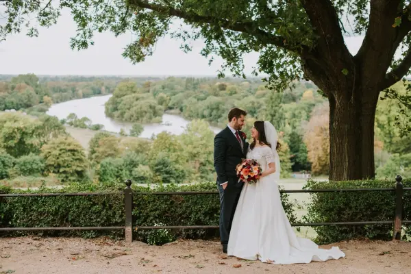Couple sharing a romantic moment by a scenic river, framed by lush greenery and a large tree, perfect for intimate outdoor weddings.