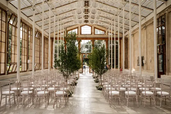 Sunlit indoor ceremony space featuring elegant Chiavari chairs lined up along a sleek aisle, surrounded by lush greenery and large glass windows.
