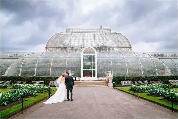 Couple embraces in front of an elegant glass conservatory, surrounded by lush greenery and charming benches for guests.