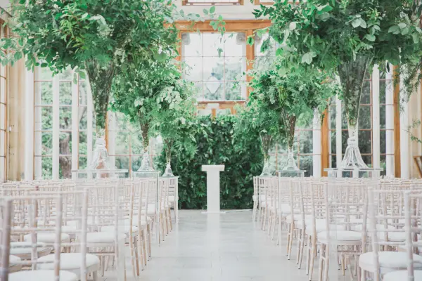 A lush indoor ceremony space adorned with greenery, elegant white chairs, and a simple altar, creating a serene and romantic atmosphere.