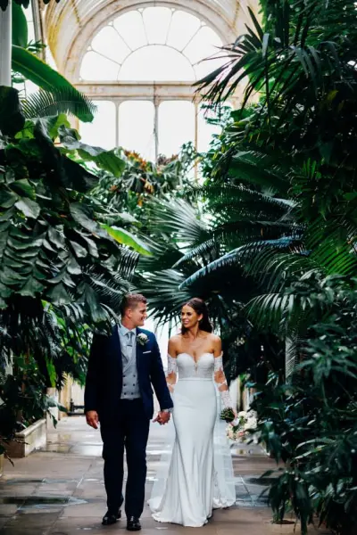 A couple strolls hand-in-hand through a lush greenhouse, surrounded by vibrant greenery and natural light streaming through large windows.