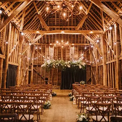 Rustic barn interior with wooden beams, elegant chandeliers, and neatly arranged chairs, adorned with floral accents for a charming wedding ceremony.
