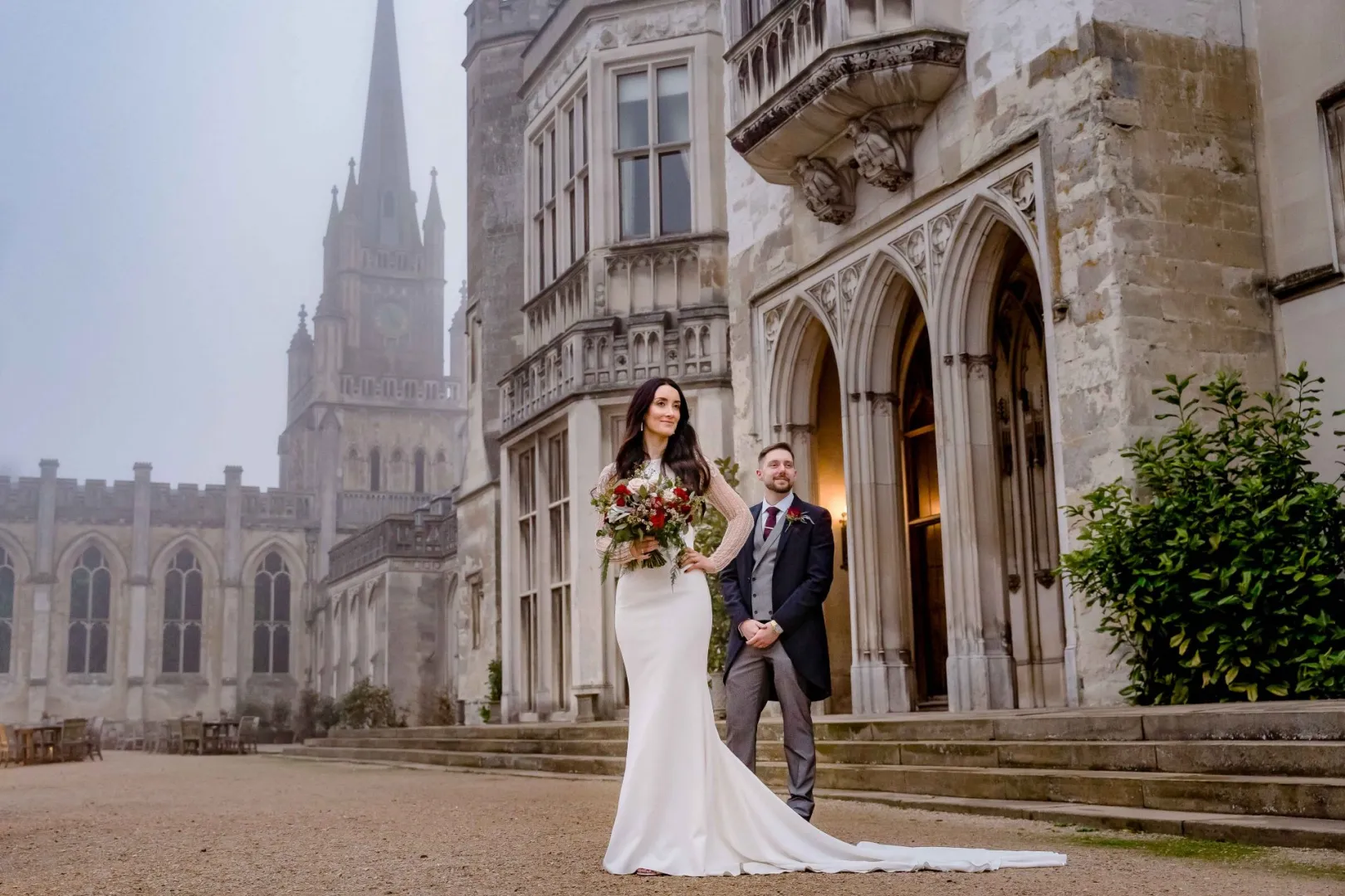 Couple stands elegantly outside a grand castle with Gothic architecture, featuring a stone facade and lush greenery nearby.