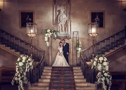 Elegant staircase adorned with floral arrangements leads to a couple standing beneath a stunning statue, creating a romantic ceremony backdrop.
