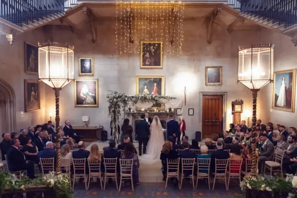 Elegant indoor ceremony featuring high ceilings, vintage artwork, and beautiful chandeliers, surrounded by guests on chic wooden chairs.