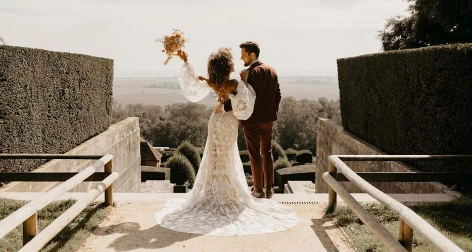 A bride in a lace gown holds a bouquet, gazing at scenic gardens alongside her partner, framed by manicured hedges.