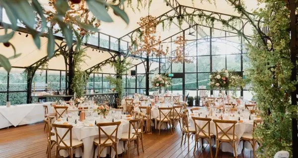 Charming glass-enclosed venue with wooden floors, elegant round tables, lush greenery, and stunning floral arrangements for a romantic celebration.