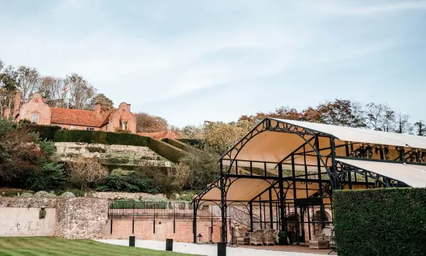 Charming outdoor venue featuring a glass pavilion, manicured gardens, and a historic backdrop of stone ruins atop a hillside.