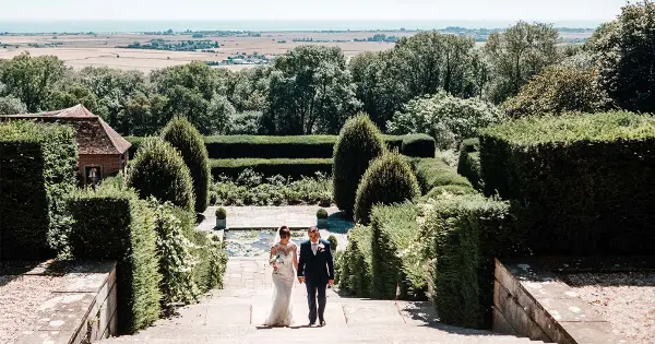 A bride and groom walk hand in hand down stone steps, surrounded by lush greenery and manicured hedges, overlooking scenic countryside.