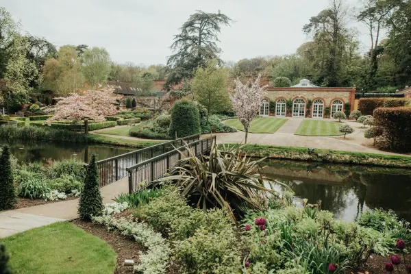 Lush gardens frame a tranquil pond, with vibrant flowers, manicured lawns, and a charming bridge leading to elegant event spaces.