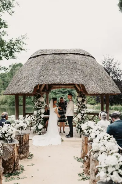 A charming outdoor ceremony under a thatched gazebo, adorned with floral arrangements and surrounded by rustic wooden seating.