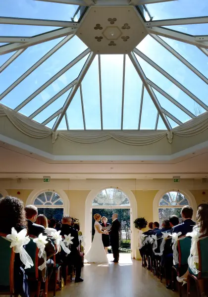 Sunlit indoor ceremony with a glass ceiling, elegant arches, and guests adorned with white floral accents witnessing the couple's vows.