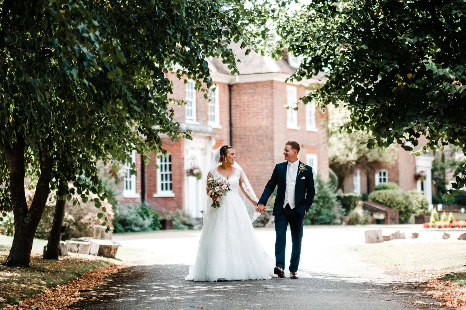 A bride and groom walk hand in hand along a tree-lined path, featuring a charming brick venue in the background. Perfect for romantic weddings.