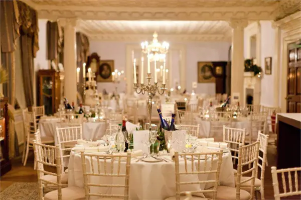 Elegant dining setup in an opulent ballroom, featuring ornate chandeliers, round tables with soft linens, and chiavari chairs.