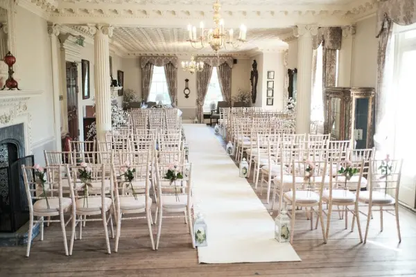 Elegant indoor ceremony space featuring rows of chiavari chairs lined with floral accents, a soft carpet aisle, and a grand chandelier.