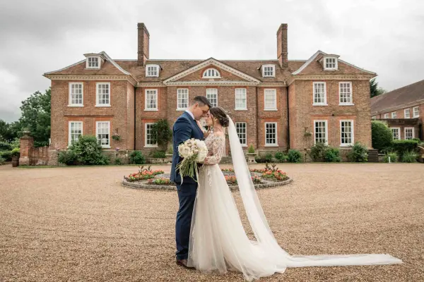 A couple shares a romantic kiss in front of a stately brick mansion, surrounded by lush gardens and a gravel courtyard.