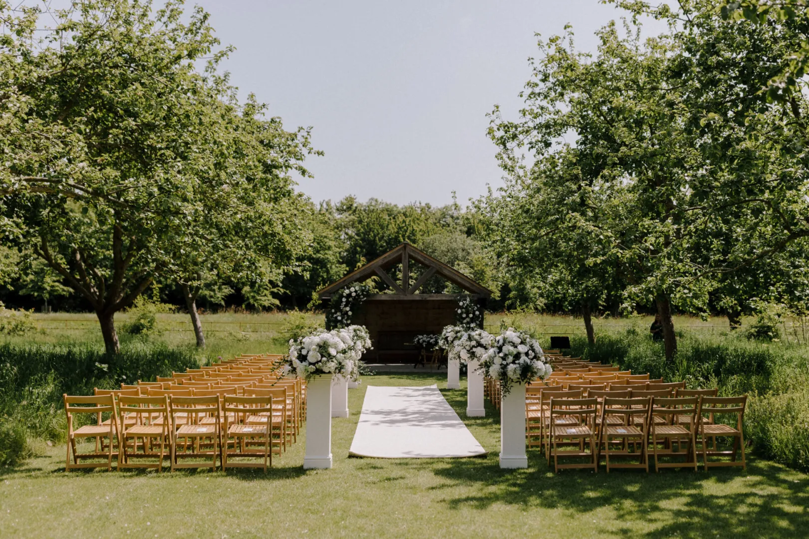 Charming outdoor ceremony space featuring wooden chairs, floral arrangements, and a lovely canopy under lush trees, set on green grass.