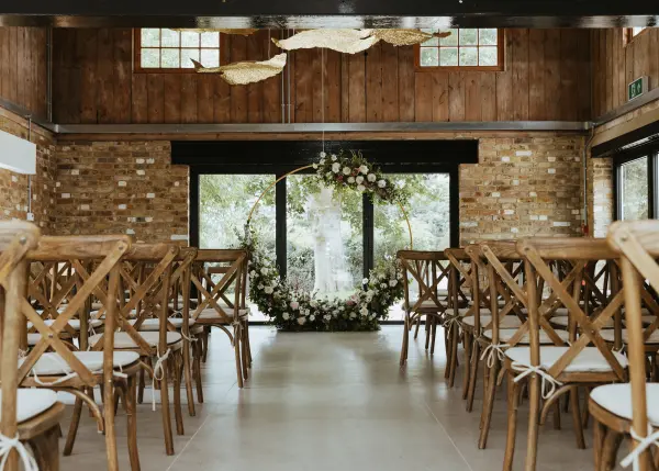 Warm wooden interiors frame a charming ceremony space, featuring crossback chairs and a floral archway by large windows.