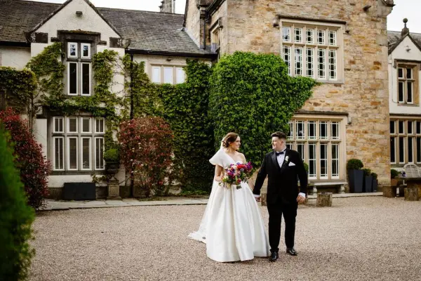 A couple strolls hand-in-hand by an ivy-covered stone building, showcasing a romantic outdoor setting perfect for weddings.