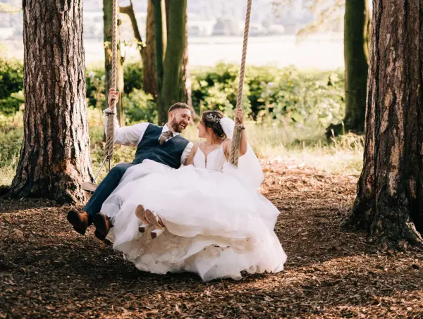 A joyful couple swings together under towering trees, surrounded by lush greenery and a serene, natural backdrop perfect for wedding photos.