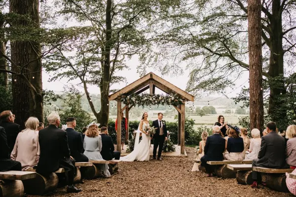 Woodland wedding ceremony space with a rustic wooden arch, surrounded by trees and seated guests on natural wooden benches.