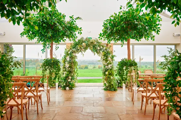 Lush greenery frames an elegant ceremony arch, with wooden chairs set against panoramic windows showcasing a scenic view.
