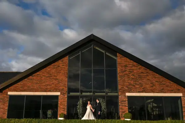 Couple standing in front of a modern brick venue with large windows, framed by lush greenery and dramatic sky.