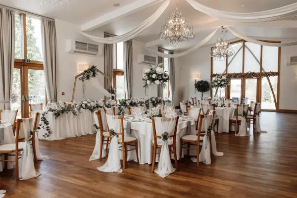 An elegant reception hall featuring wooden floors, draped chandeliers, and beautifully decorated tables with floral centerpieces.