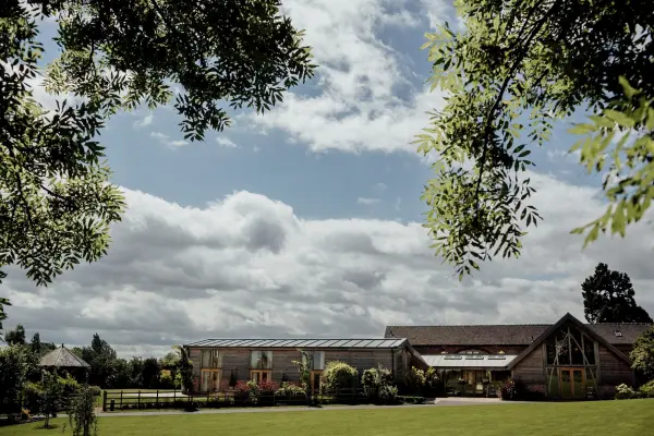 Charming countryside venue featuring a spacious lawn, rustic barn, and modern glass-enclosed area, surrounded by lush greenery and blue skies.