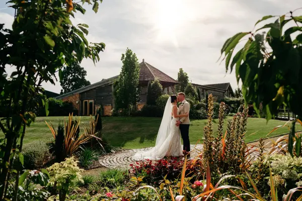 A couple shares a romantic kiss surrounded by lush gardens and vibrant flowers, with a charming venue in the background.