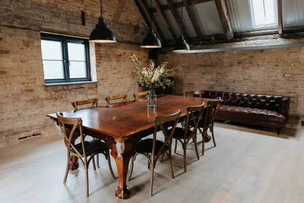 Rustic event space featuring a large wooden table, mismatched chairs, and a cozy leather sofa under exposed beams and pendant lights.