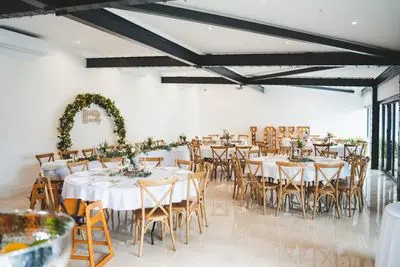 Bright indoor wedding venue featuring round tables, rustic chairs, and a green floral arch, with sleek beams and large windows for natural light.