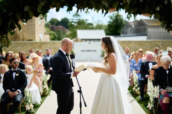 A beautiful outdoor ceremony space featuring a floral arch, with the couple exchanging vows surrounded by seated guests and lush greenery.