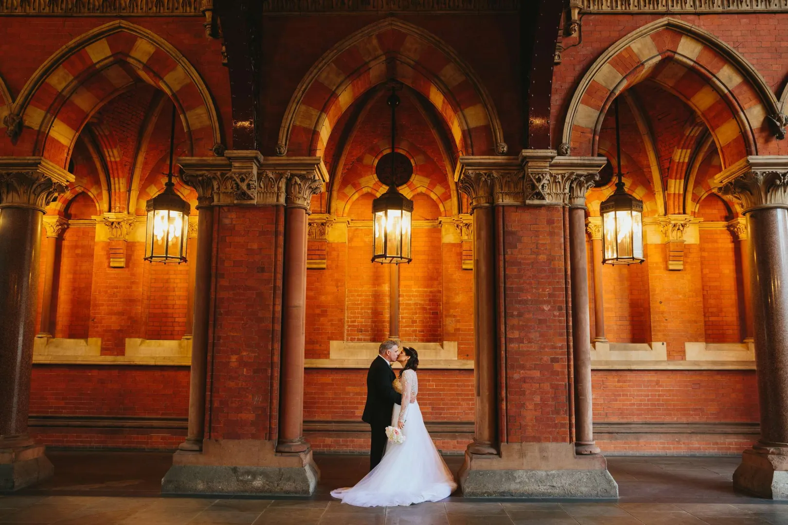 Couple embraces inside a grand hall featuring stunning arched ceilings, warm brick walls, and elegant lanterns for a romantic atmosphere.