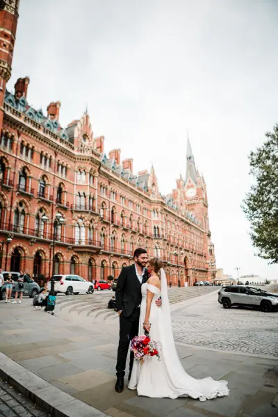 Couple embraces in front of a stunning red-brick Victorian building, perfect for a romantic wedding backdrop. Elegant architecture and vibrant flowers.