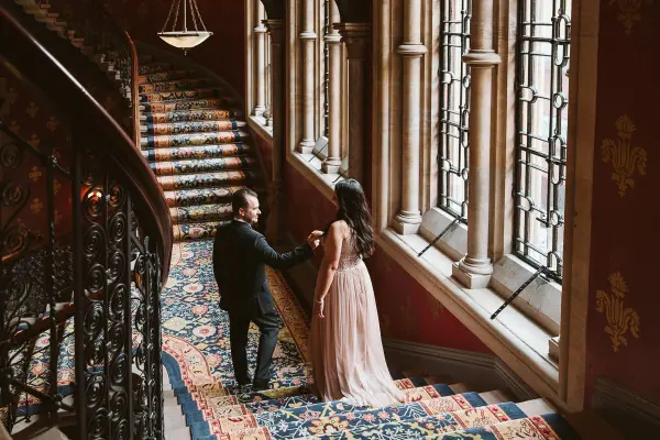 Elegant couple ascending a grand staircase adorned with rich carpets and large windows, creating a romantic atmosphere for a wedding.