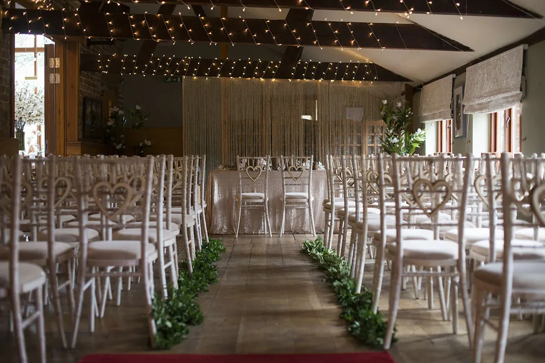 Charming indoor ceremony space with heart-back chairs, twinkling lights, and greenery lining the aisle, creating a romantic atmosphere.