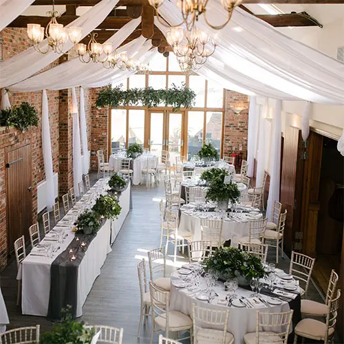 Elegant indoor reception space featuring long tables adorned with greenery, exposed brick walls, and chandelier lighting. Perfect for a wedding.